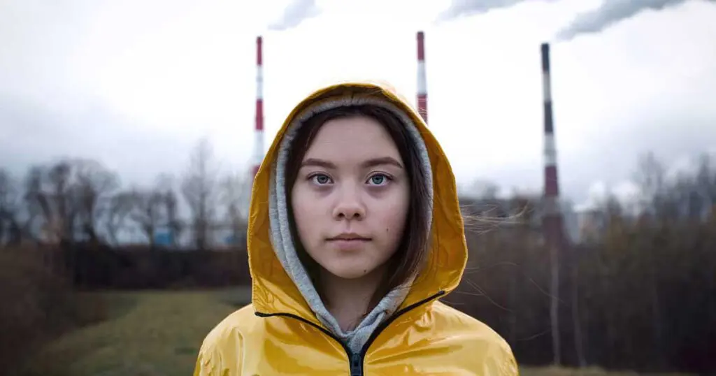 Young person in a yellow jacket stares into the camera as three smoke stacks billow into a graying sky from a plume in the distance.