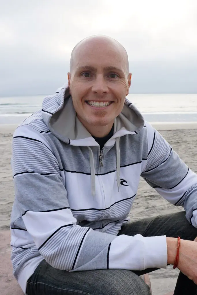 Adam Footer smiles while sitting on a beach in his striped hoodie, the ocean shimmering in the background.