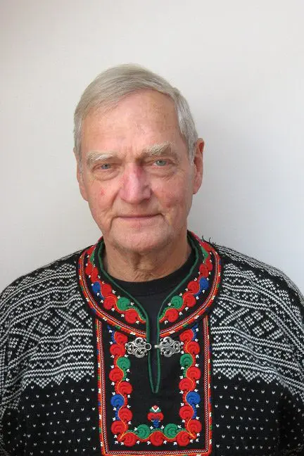 An elderly man with gray hair wears a traditional knit sweater adorned with colorful patterns and a metal clasp, standing against a plain white background.