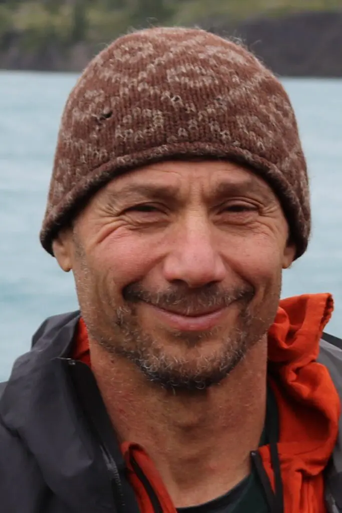 A man with a beard and mustache, identified as author Alejandro Frid, is wearing a brown knit hat and a black and orange jacket. He is smiling with a body of water and greenery in the blurred background.