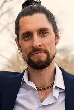 Blake Cothron, a man with a beard and tied-back hair, exudes sophistication in his suit and necklace while enjoying the outdoors.