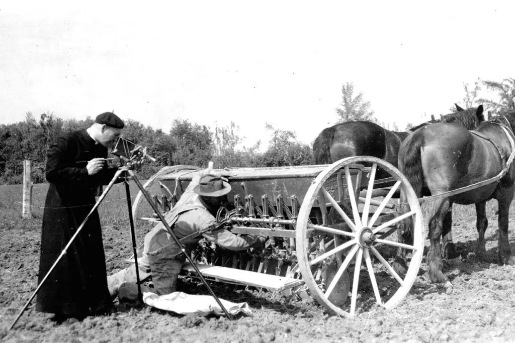 Black and white photo of farmer fixing his horse-drawn plow as a photographer takes a photo.