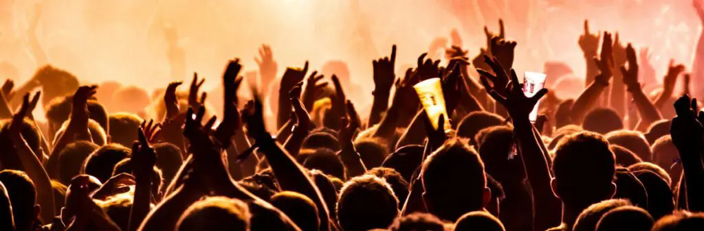 Concert crowd with hands raised, illuminated by warm, vibrant lighting. Some people hold cups.