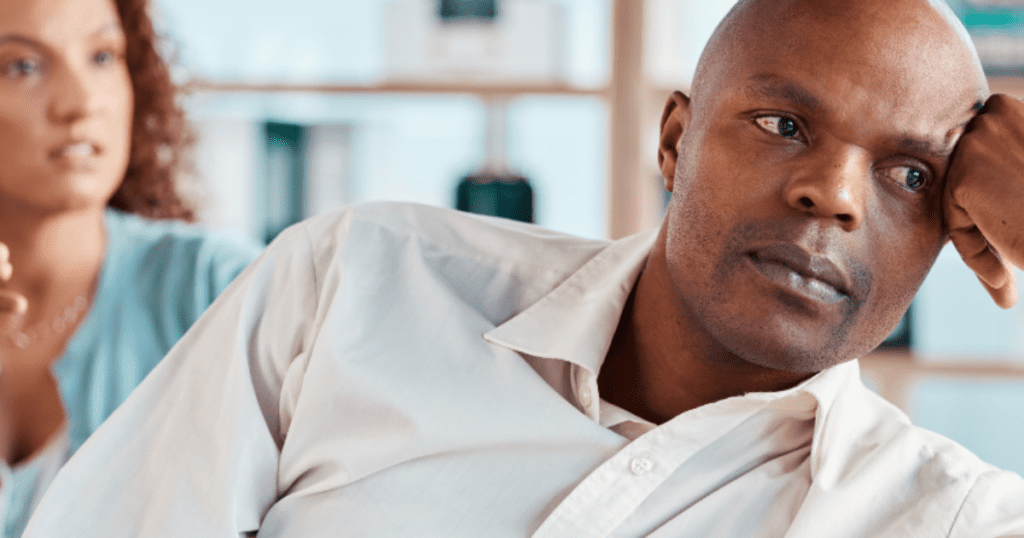 A man leans away from a woman staring at him angrily while sitting on a couch.