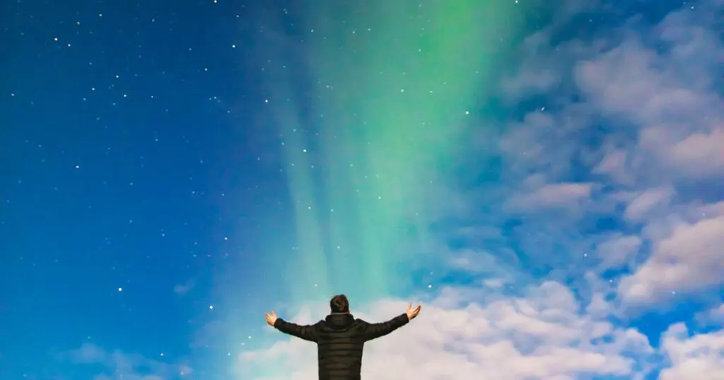 A person stands with arms outstretched under a night sky illuminated by the Northern Lights, featuring shades of green and blue against a backdrop of stars and clouds. The individual appears to be wearing a dark coat and is facing the sky, evoking a sense of awe and wonder suitable for the scene described in Social_Excerpt_Introduction-to-Power.