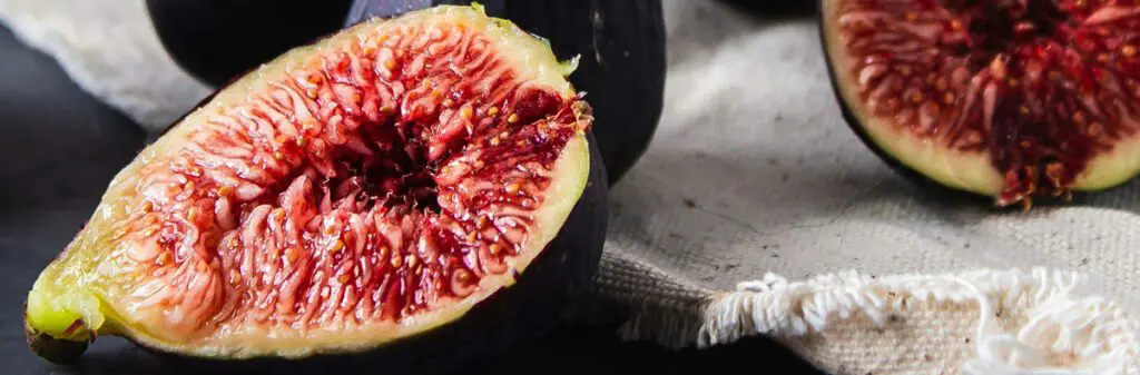 Close-up image of several ripe figs with a focus on a halved fig displaying its vibrant, juicy, and seed-filled interior. The figs are placed on a rustic, textured white cloth, with more whole figs visible in the background, creating an inviting, fresh display.