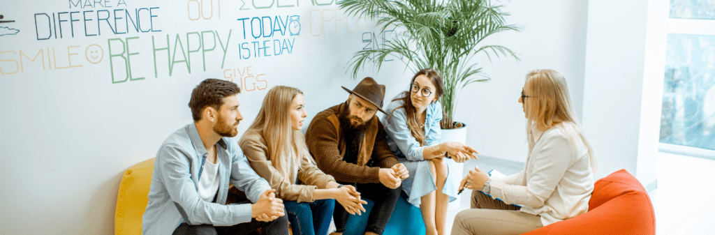 A group of young adults sit in front of a wall featuring positive affirmations, having a discussion with a leader or counselor.