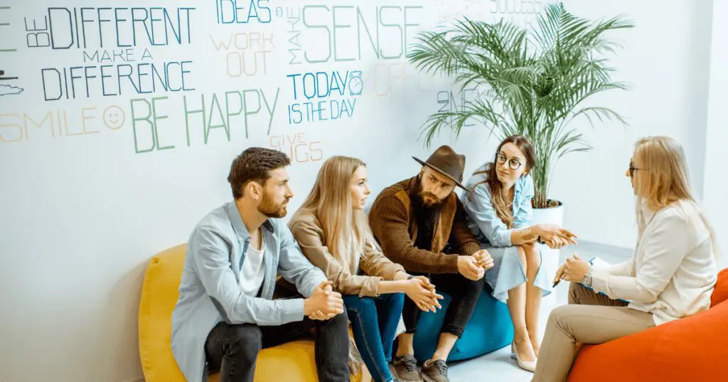 A group of young adults sit in front of a wall featuring positive affirmations, having a discussion with a leader or counselor.