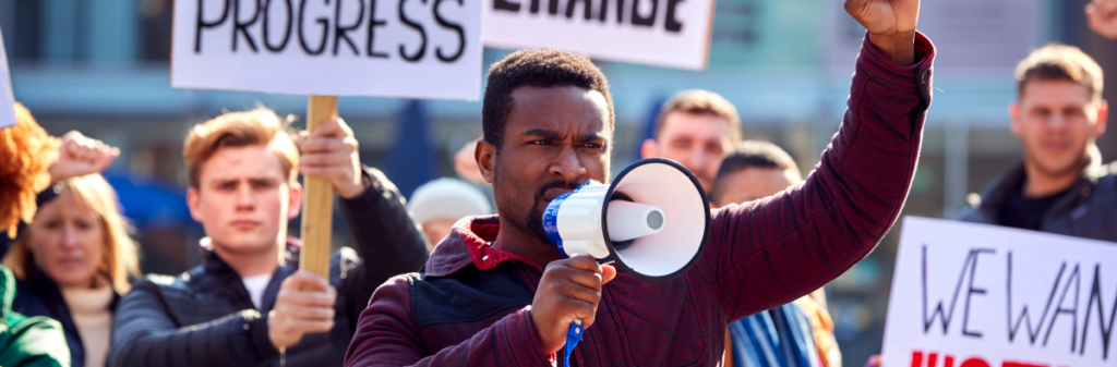 A diverse group of people protests outdoors, featuring the focal individual, who is holding a megaphone and raising their fist.