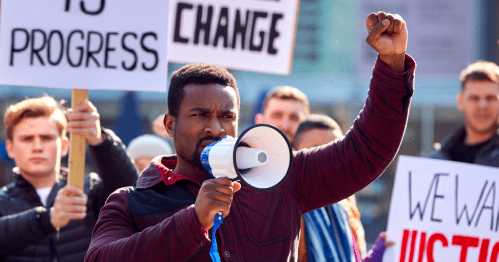 A person stands at the forefront of a protest, holding a megaphone and raising their fist. Protesters in the background hold signs with messages such as "Progress" and "Change." The atmosphere is spirited and determined, perfectly illustrating the ethos captured by NVC_Exit_Social-Image.