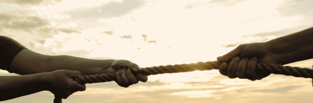 Two people engage in a game of tug-of-war, each pulling on opposite ends of a rope. The sun sets in the background, casting a warm glow over their hands.