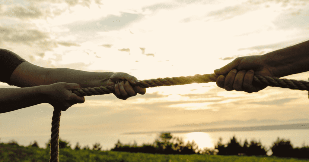 Two pairs of hands gripping a rope in a tug-of-war during sunset with a scenic landscape in the background.