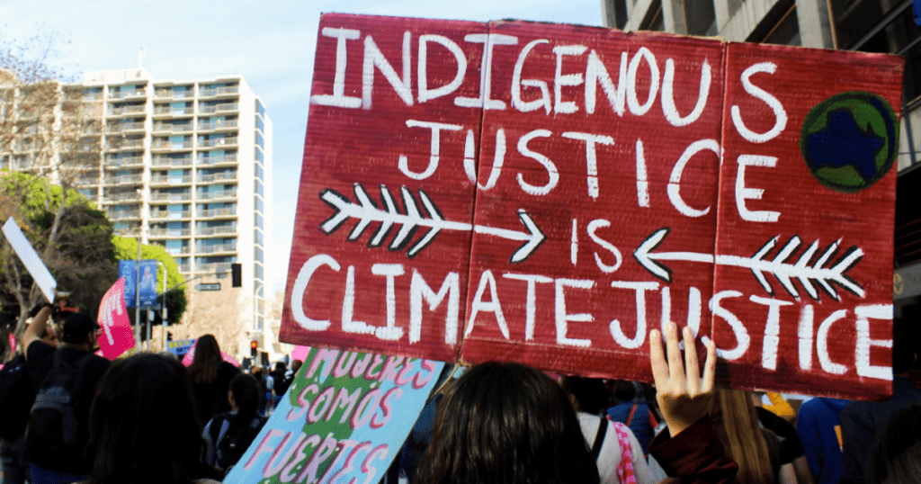 A protest sign reads "Indigenous Justice is Climate Justice" with arrows and a globe, emphasizing the interconnectedness of Climate Action on the National Day for Truth and Reconciliation.