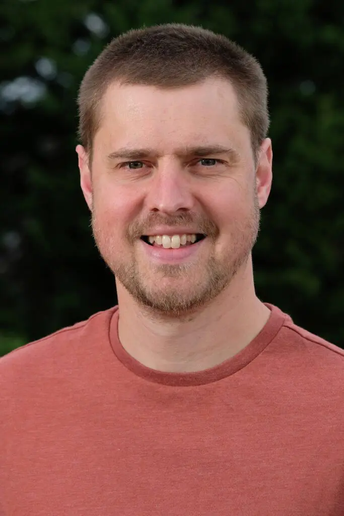 Trevor Piersol, a man with short hair and a beard, smiles while wearing a reddish-brown shirt, with trees in the background.