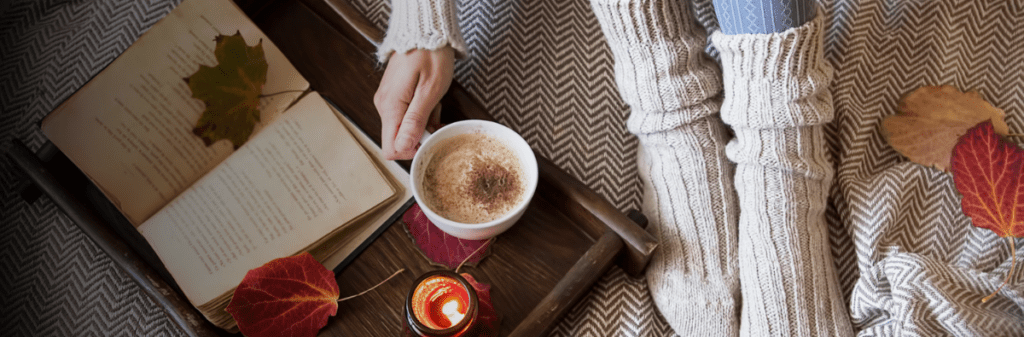 In a cozy setting perfect for conscious gifting, a book rests alongside warm socks, a candle, and a cup of coffee on a tray adorned with fall leaves.