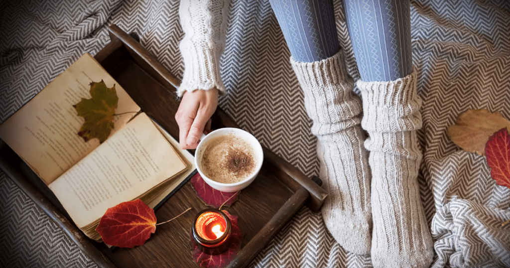 A cozy scene unfolds with a person holding a coffee on a tray, accompanied by a book, candle, and autumn leaves, embodying the essence of thoughtful gifts and conscious gifting.