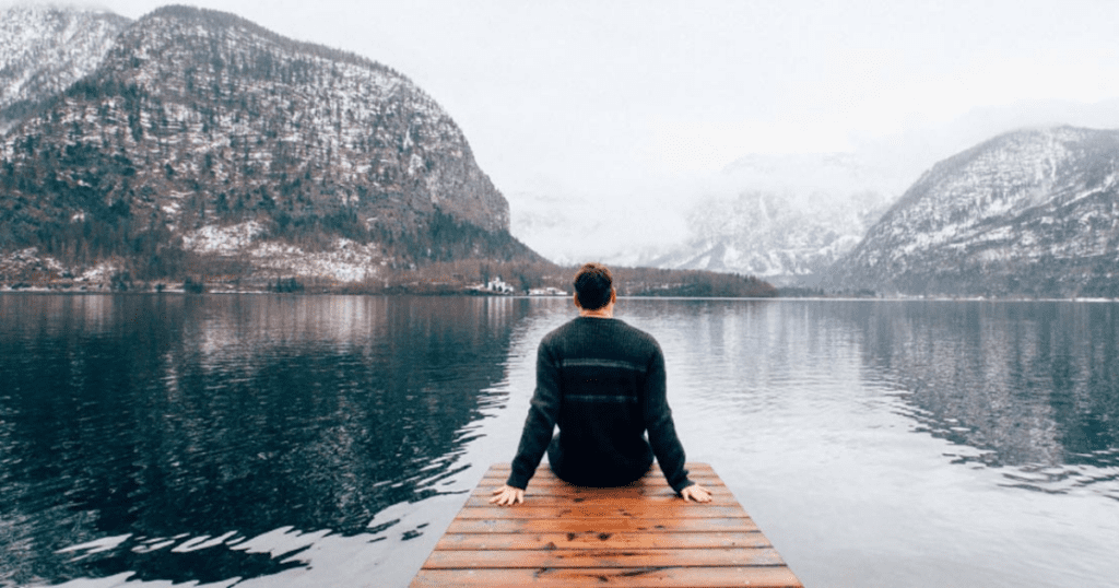 A person sits on a wooden dock, reflecting on how change actually happens while overlooking a tranquil lake and snow-capped mountains.