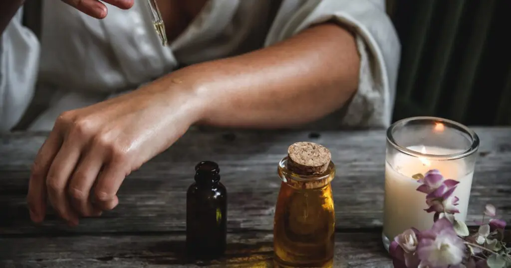 A person is carefully applying handmade oil to their arm, the gentle glow of a candle casting warm shadows over the wooden table adorned with charming bottles. This serene moment feels like a holiday for the senses.