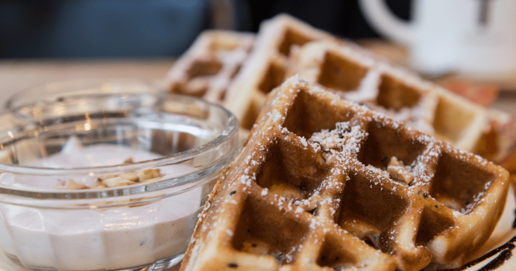 Close-up of zucchini waffles dusted with powdered sugar, served with a small bowl of yogurt and granola. Savor this delightful treat that transforms any breakfast into a holiday feast.