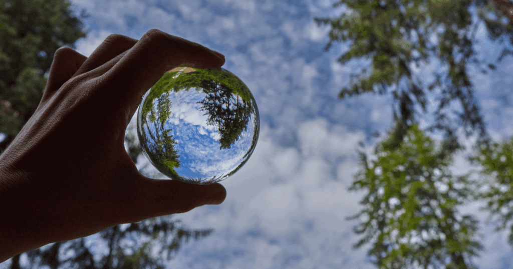 A hand holds a glass sphere reflecting trees, a cloudy sky, and an inner landscape.