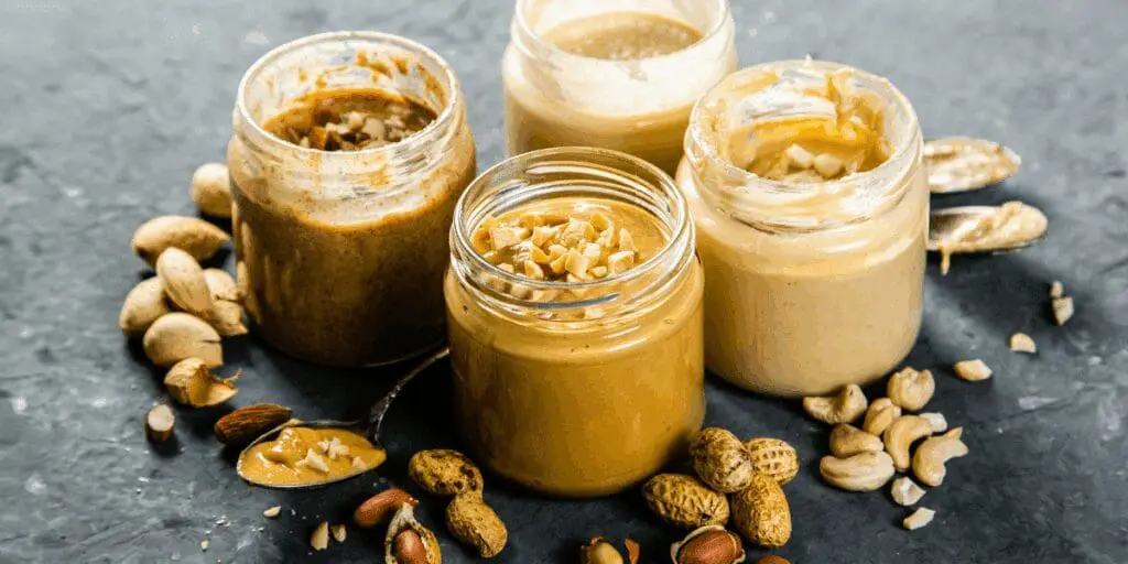 Four jars of nut butters with scattered nuts, staples of the modern American diet, on a dark surface.