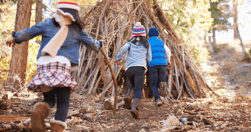 Three children in fall clothes run toward a large stick teepee, sparking nature connection.