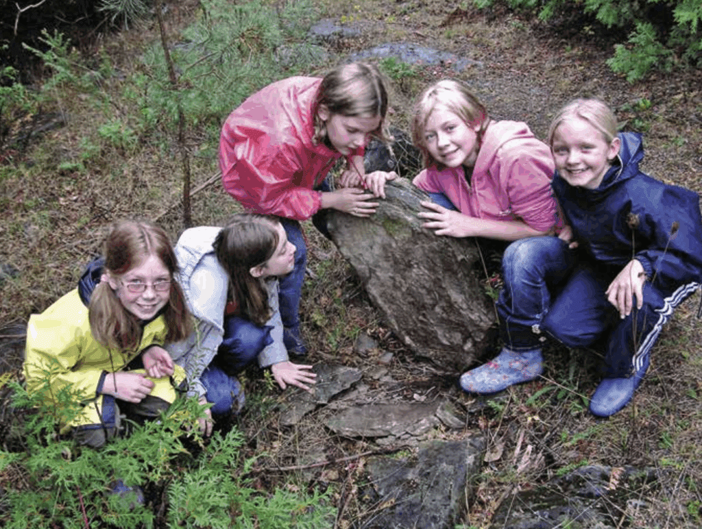 Five kids enjoy fun activities outdoors, gathered around a large flat rock and smiling brightly.