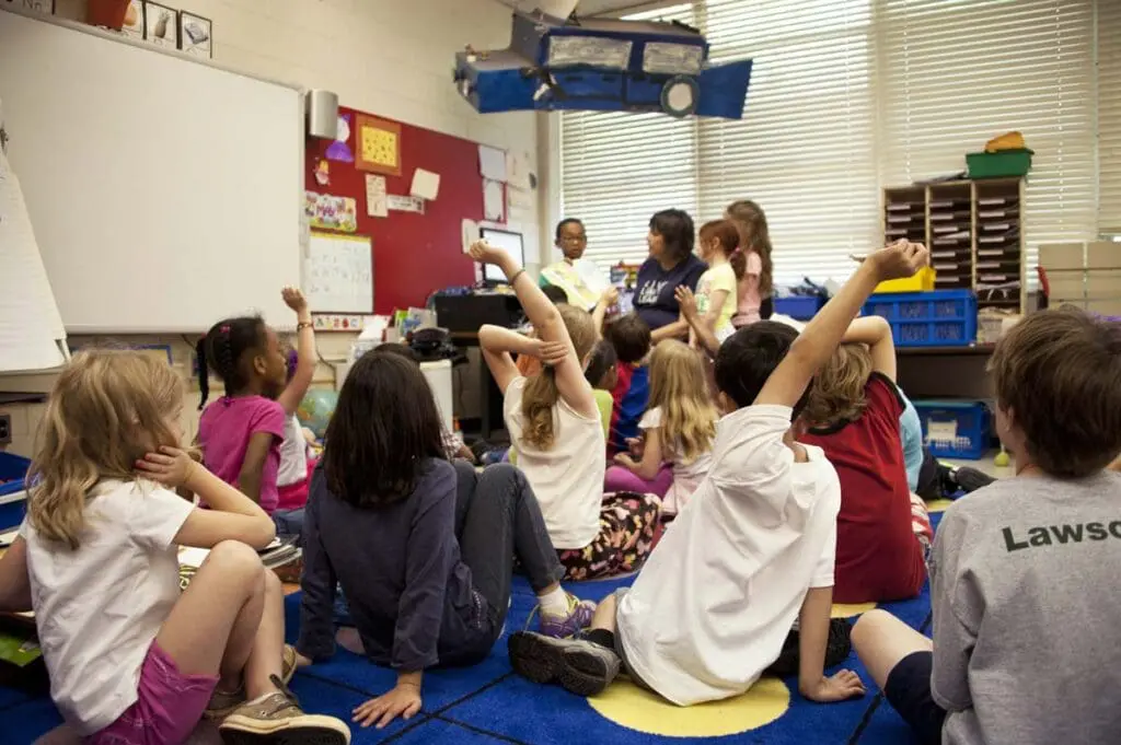 Children sit on a classroom floor, raising hands as the teacher reads from When Kids Rule the School.