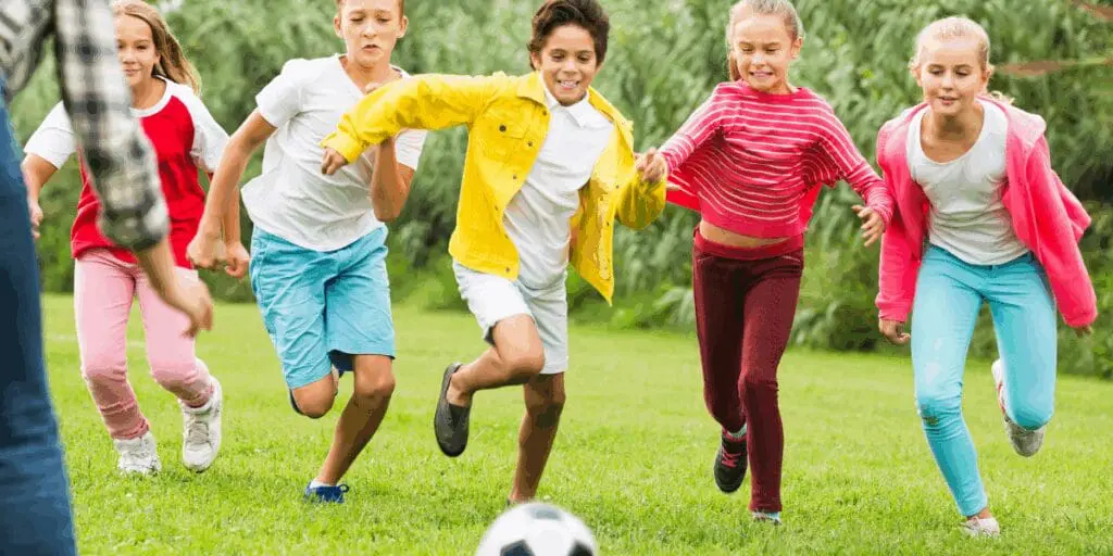 Back to school, kids chase a soccer ball on a grassy field, smiling and running with summer joy.