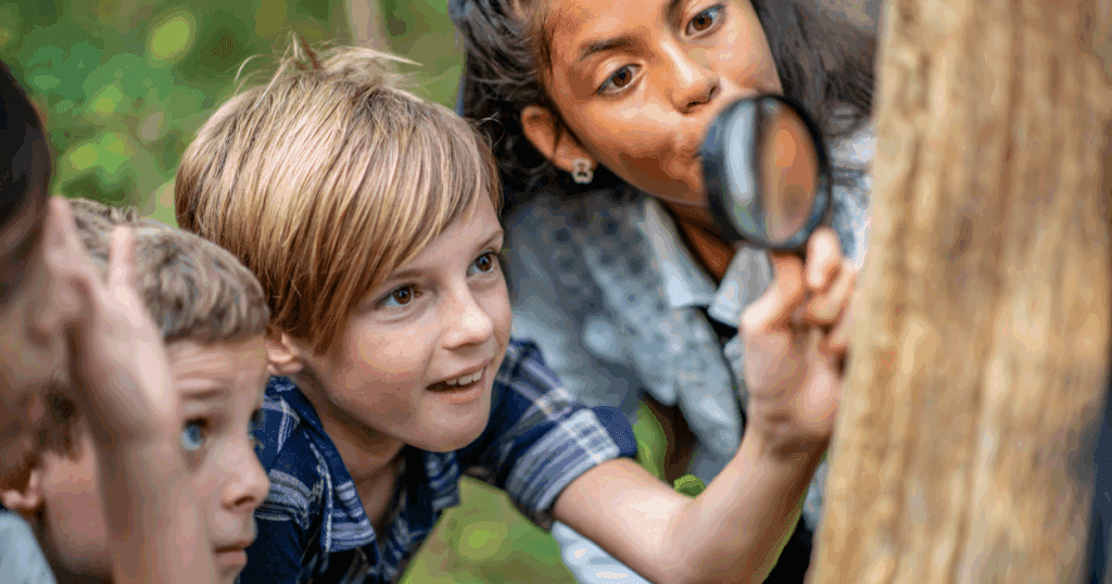 Children engaged in outdoor play, closely examining a tree with a magnifying glass.
