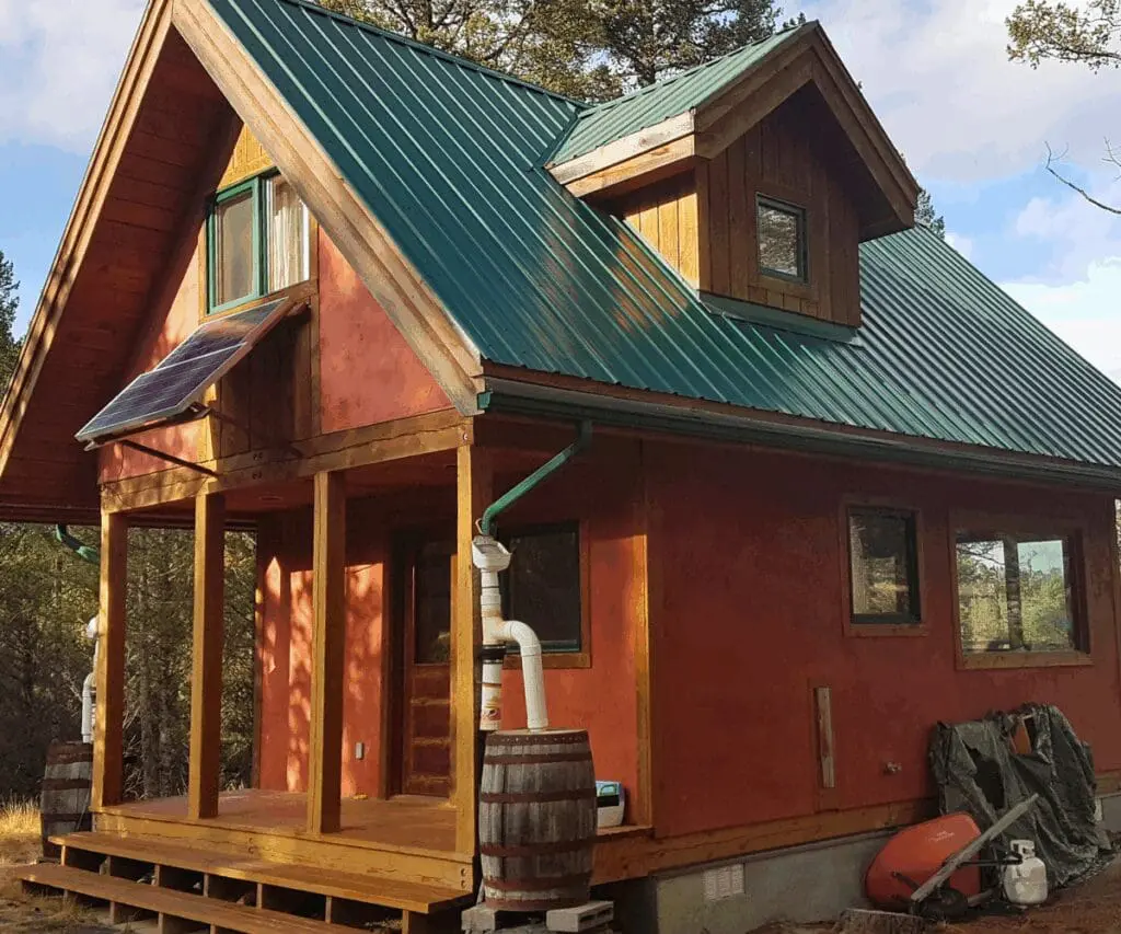 Small red cabin with green roof, porch, solar panel, and barrels for rainwater harvesting.