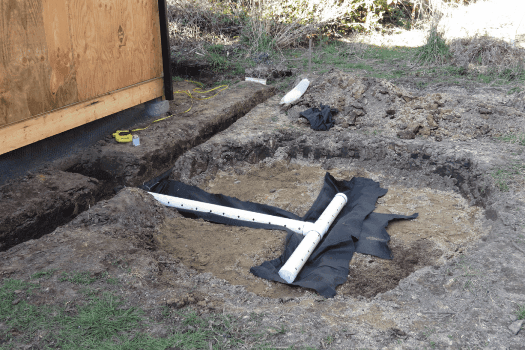 White perforated pipes and black fabric installed in a shallow dug area near a wooden shed.