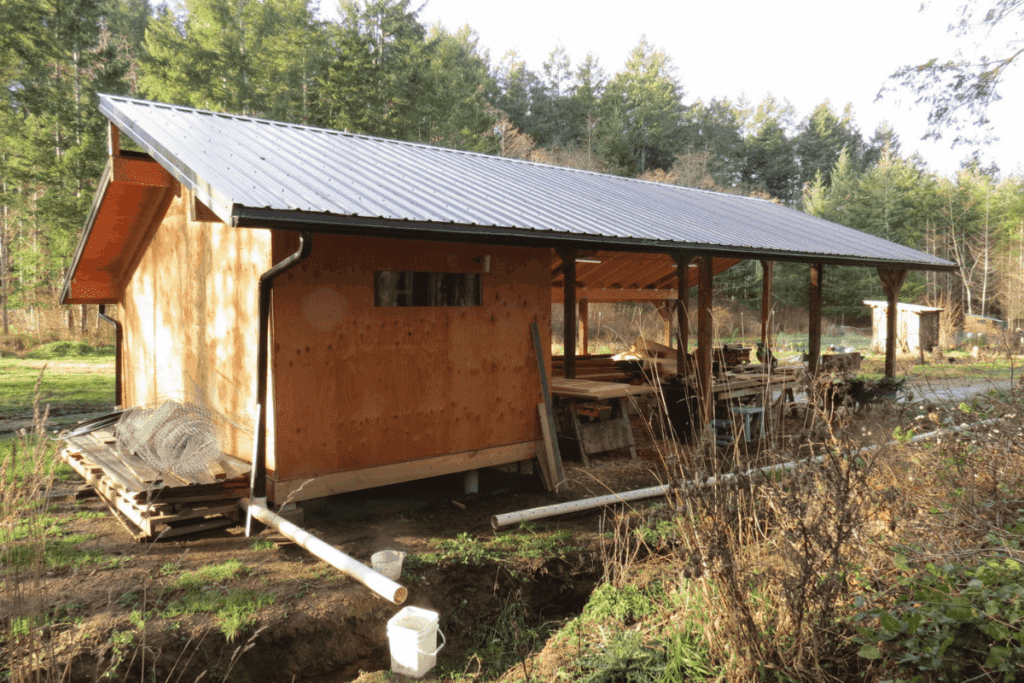 A wooden shed with a metal roof, surrounded by trees and stacked lumber outdoors.