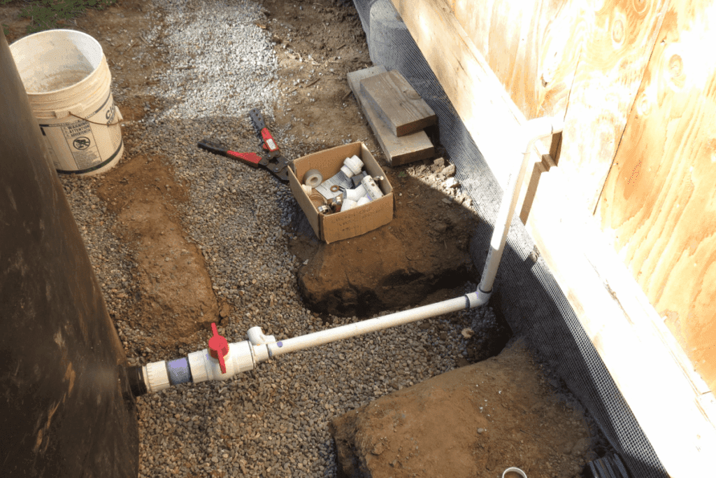 White PVC pipes with a red valve are being installed next to a building; tools and parts nearby.