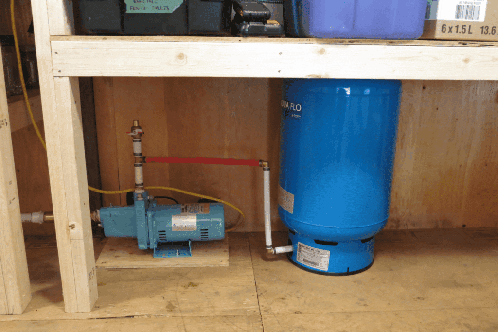 Small water pump and blue pressure tank installed under a wooden shelf in a utility area.