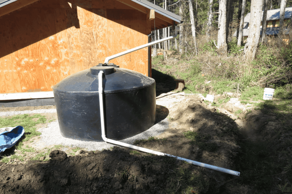 Black rainwater tank with white pipes next to a wooden building in a forested area.