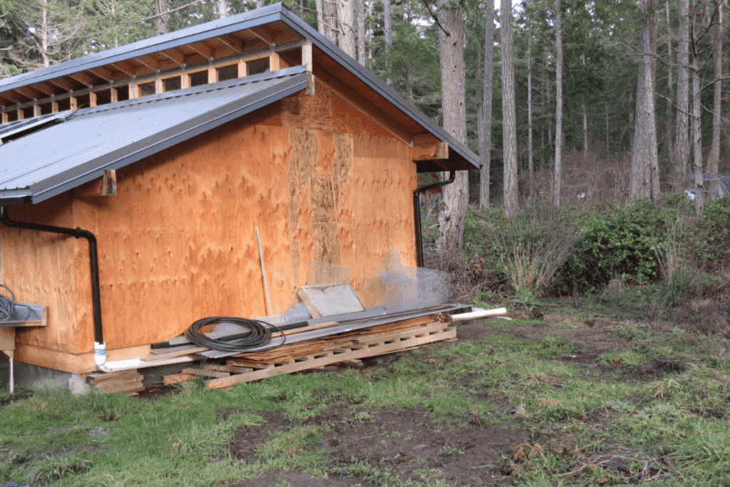A wooden shed with construction materials stacked on a pallet beside it in a forested area.