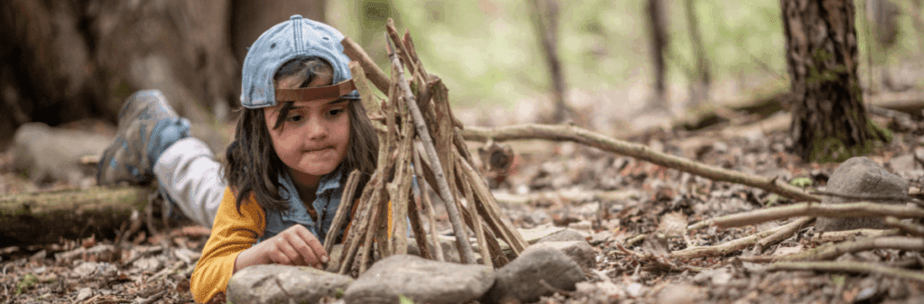 A child in a cap builds a small teepee with sticks in the forest. Outdoor play.