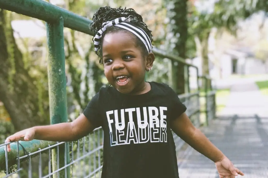 Smiling young girl in "Future Leader 2025" t-shirt stands outdoors on a walkway.