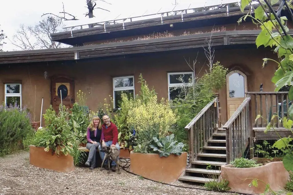 Two people and a dog relax by a garden, earth-toned house, and composting toilets on the porch.