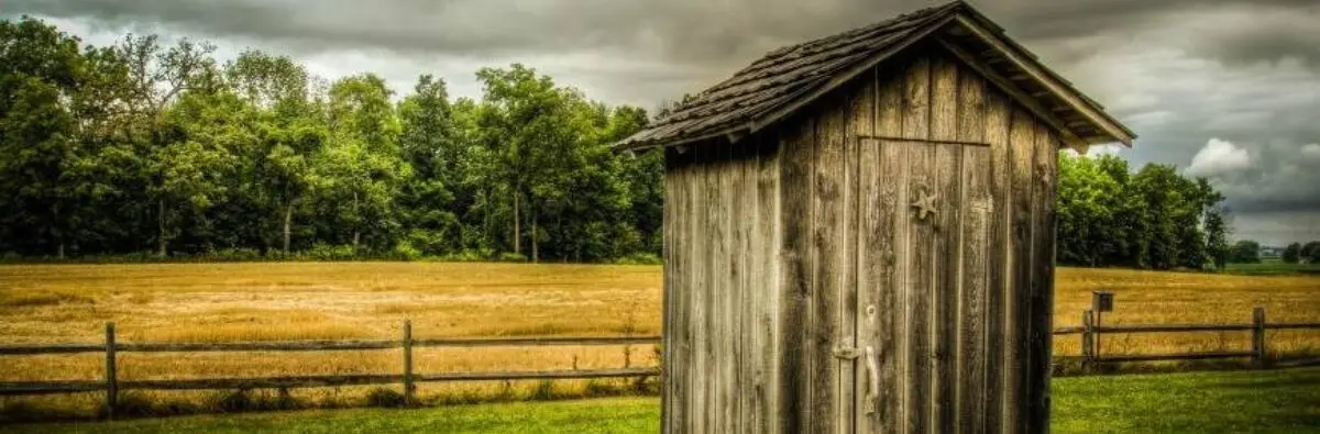 Old wooden outhouse stands in a grassy field, recalling Essential Composting Toilets design.
