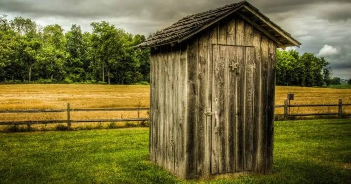 A weathered wooden outhouse stands on grass, reflecting Gord Baird’s composting toilets ethos.