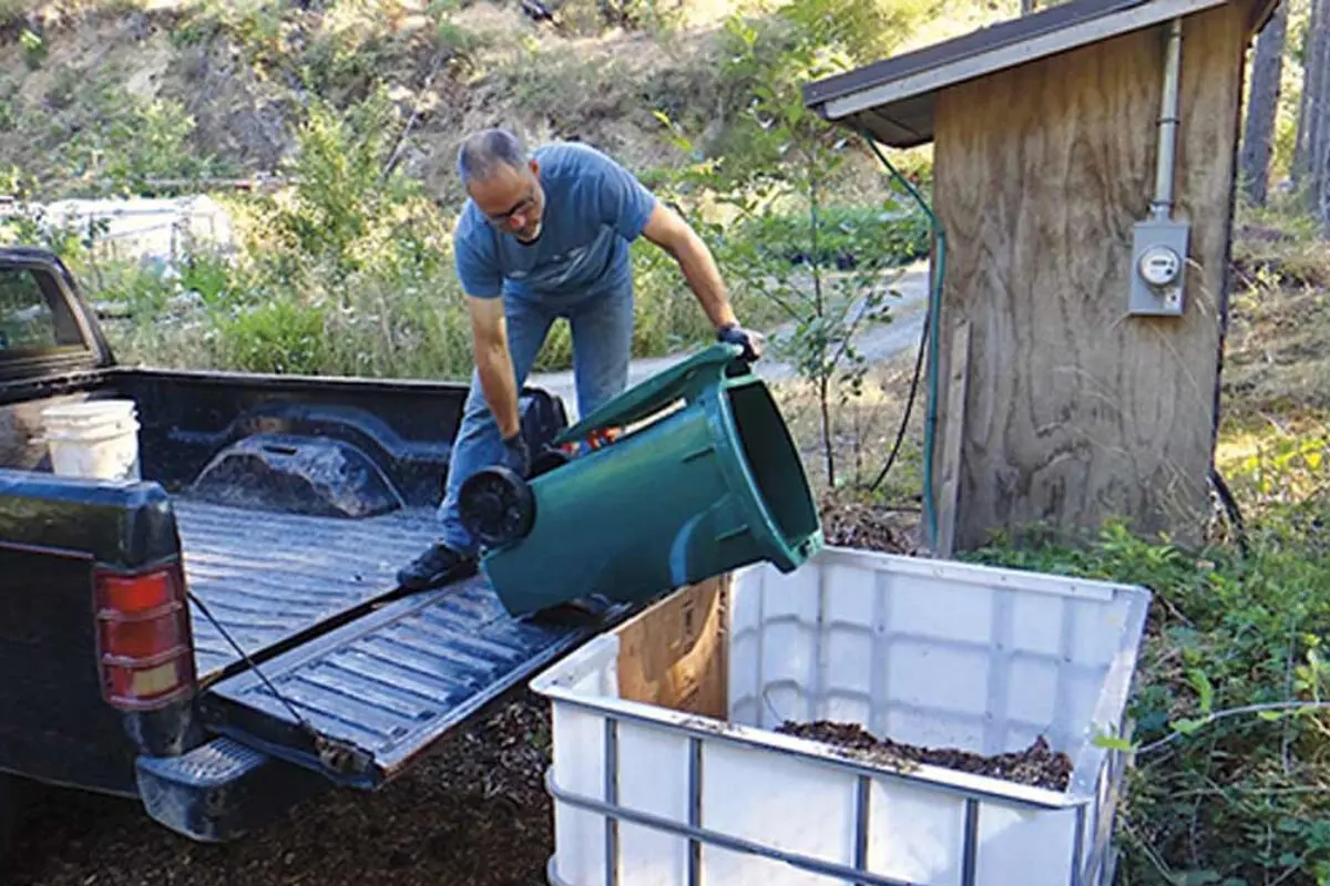 Man empties green bin of organic waste into large white container beside composting toilets.