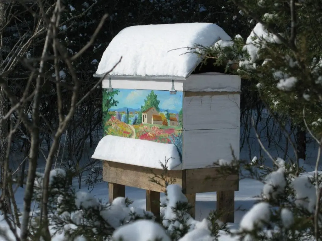 A bee-friendly, snow-covered beehive with a colorful painting, nestled among trees.