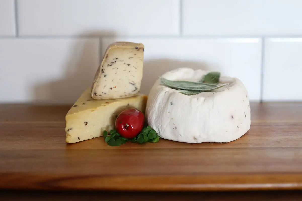 Three types of homemade cheese with herbs, tomato, and parsley on a board against white tile.