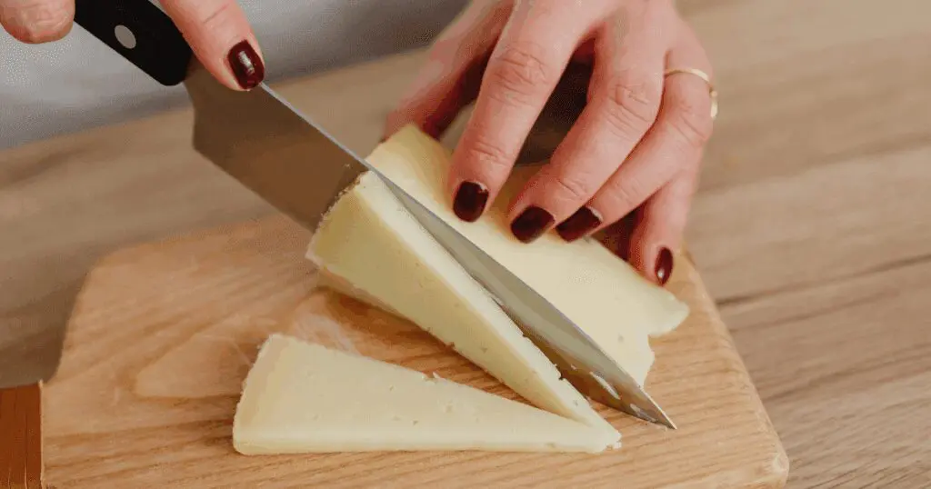 A person slicing homemade cheese into triangles on a wooden cutting board with a large knife.