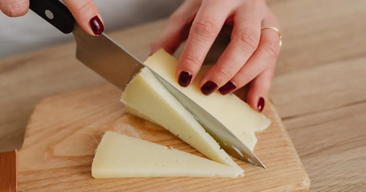 A person slicing homemade cheese into triangles on a wooden cutting board with a large knife.