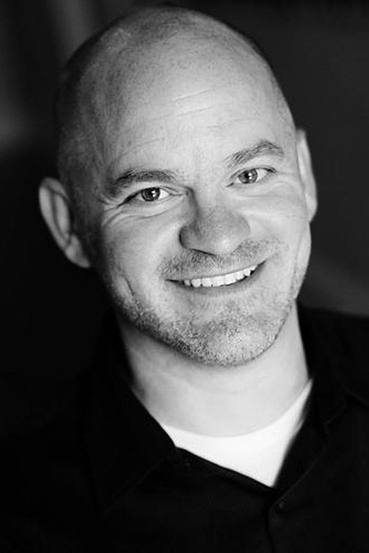 Black-and-white portrait of a smiling bald man with light facial hair, wearing a dark shirt over a white undershirt, looking directly at the camera—author_Aaron-Lubeck.