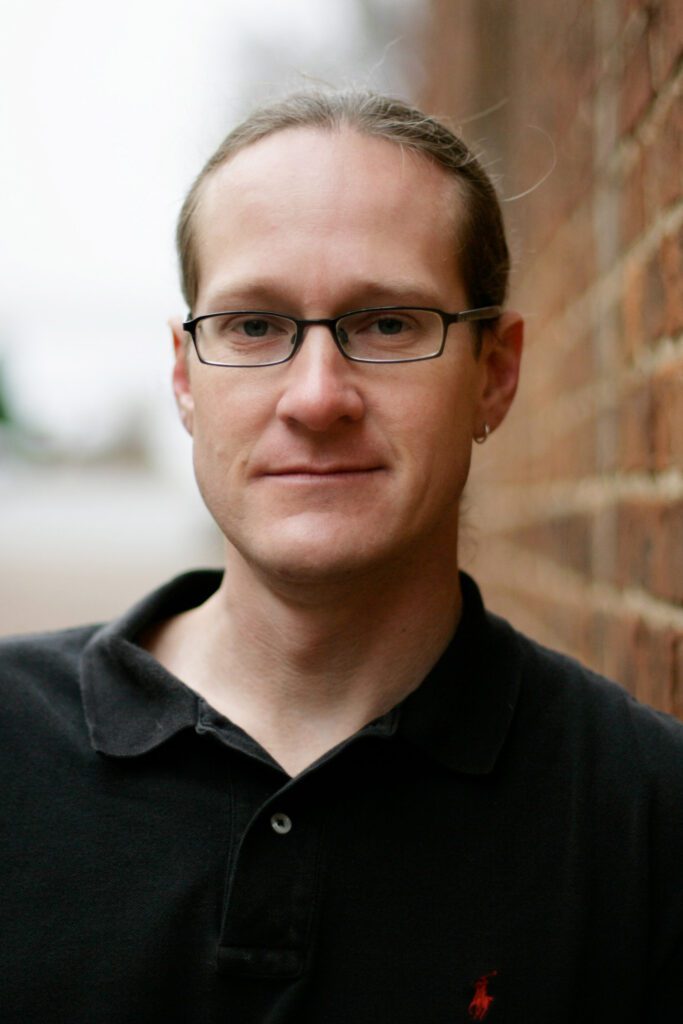 author_Aaron-Newton, a man with glasses, wearing a black polo shirt and small hoop earring, stands against a brick wall.