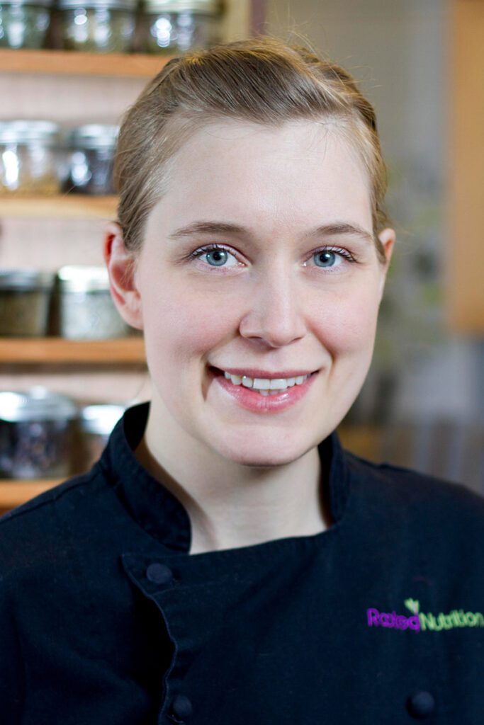 Andrea Potter, with light hair and blue eyes, stands in a black chef's coat featuring an embroidered logo against a backdrop of shelves filled with jars.